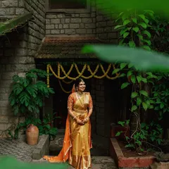 A bride in a golden saree, framed by the doorway of a stone cottage and lush greenery, smiles, bathed in soft, natural light.
