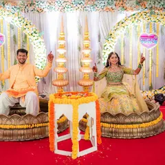 The happy couple at their Haldi ceremony. The backdrop features large floral rings and traditional brass lamps, perfectly complementing the joyous occasion.