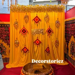 A classic Haldi decoration featuring a bright yellow backdrop, which is a traditional color for the ceremony. The setup is adorned with handcrafted hangings, a 'हळद' sign, and a large brass Urli decorated with marigold flowers.