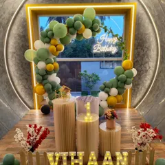 A modern and minimalist setup with an olive green and yellow balloon arch framing a window. A neon sign and marquee letters add a warm glow.