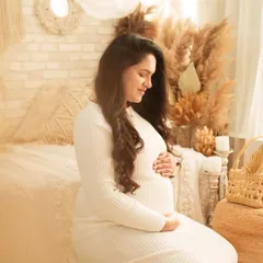 A quiet moment of reflection for a mother-to-be in a cozy boho-chic room. Dressed in a comfortable white ribbed dress, she kneels and cradles her bump, surrounded by pampas grass and soft textures.
