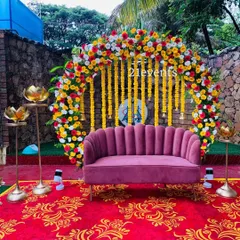 A vibrant and colorful setup perfect for a Mehendi or Haldi ceremony. The backdrop features a floral arch of marigolds and roses with hanging yellow flower strings, set against a modern pink sofa for a fun, festive photo opportunity.