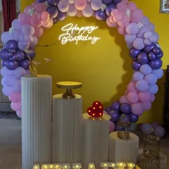 A stylish birthday setup with a circular balloon arch in shades of purple and pink. The decor is completed with a 'Happy Birthday' neon sign and modern white plinths.