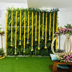 An elegant naming ceremony setup with a modern touch. The backdrop is a wall of green leaves and hanging marigold strings, while the circular wooden cradle is adorned with fresh pink roses.