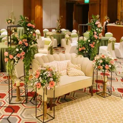 We created a sophisticated lounge area for a milestone birthday party. A plush scalloped sofa is surrounded by modern black stands holding beautiful arrangements of pink and white flowers, creating a perfect photo spot for guests.