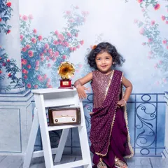 This little girl looks so poised and confident in her lovely purple pre-stitched saree. The vintage props like the radio and gramophone add a touch of old-world charm to the portrait.