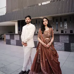 A full-length shot of Piyush and Anjali, showcasing their coordinated engagement outfits. The groom is in a sophisticated white sherwani, and the bride wears a beautiful brown embellished lehenga.