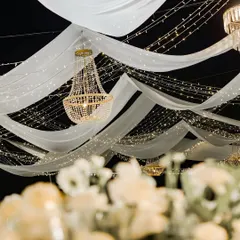 I use lighting and fabric to completely transform a venue. Here, flowing white drapes, strings of fairy lights, and classic crystal chandeliers create a dreamy and romantic ceiling for a reception dinner.