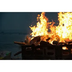 A close-up of a funeral pyre with the Ganga river in the background. The focus here is on the raw power of the fire itself, an element that is both destructive and purifying in Hindu tradition.