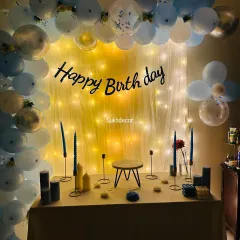 A classic birthday party setup with a 'Happy Birthday' sign against a curtain of fairy lights. The scene is framed by a balloon arch in shades of blue, white, and silver, with a table set for cake and gifts.