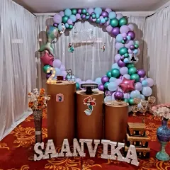 A mermaid theme is always a favorite for kids' birthdays. This setup for Saanvika features a circular balloon arch in ocean colors, themed cutouts of Ariel, and pedestals for the cake and gifts.