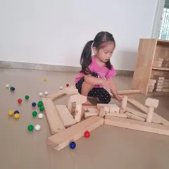 A child creates a long, winding path with wooden blocks and colorful marbles. This is a self-directed experiment in physics and design, as she figures out how to make the marbles roll.