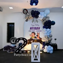 A "Two Fast, 2 Curious" car-themed party for a second birthday. The decoration includes a large number '2' shaped like a road, a personalized backdrop, and a blue, black, and grey balloon arch.