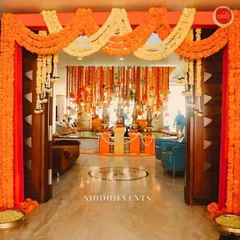 A classic Haldi entrance, rich with tradition and color. The doorway is framed by thick garlands of orange and yellow marigolds, leading into a space filled with hanging floral strings, creating a warm and auspicious welcome for all.