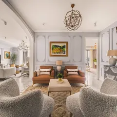 This is a perspective of a neoclassical living room, showcasing a masterful blend of classic elegance and modern comfort. The plush, textured armchairs in the foreground invite you into a space defined by symmetrical design, ornate wall moldings, and a grand, gilded mirror that amplifies the room's luxurious feel.