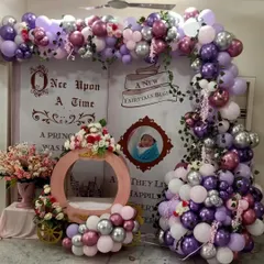 This "Once Upon a Time" setup was designed for a baby girl's welcome home party. It features a custom fairytale book backdrop, a princess carriage prop for photos, and a lush balloon garland in shades of purple, pink, and silver.
