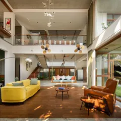 The main living area is conceived as a pavilion, with a double-height ceiling and large openings to the garden. The space combines natural materials like wood and stone with modern comforts, centered around a traditional Indian swing.