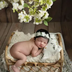 A peaceful newborn rests in a simple wicker basket, adorned with a delicate floral headband. The natural textures and soft lighting highlight the baby's serene expression and tiny features.