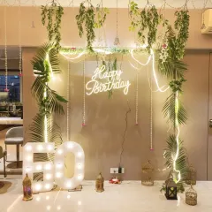 A tropical-themed 50th birthday decoration. This fresh look combines a 'Happy Birthday' neon sign with lush greenery, hanging floral elements, and large marquee numbers.