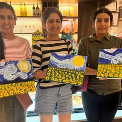 A trio of friends proudly displaying their textured sunflower and sky paintings. These workshops are a perfect creative hangout.