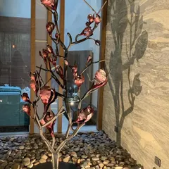A sculptural steel tree with copper-toned leaves sits in a bed of river pebbles inside a home. This piece of art and its shadow play on the marble wall behind it are an example of how I bring elements of nature indoors in an abstract and artistic way.