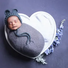An overhead shot of a baby in a bear-eared hat, nestled in a heart-shaped bowl. The simple grey and blue color palette creates a calm and sweet image.