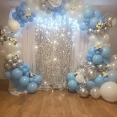 A beautiful balloon arch in a 'Frozen' theme, using shades of blue, silver, and white. The sparkling curtain in the middle adds a touch of magic to this home party setup.