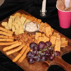 A close-up of a rustic wooden grazing board, perfect for any party. This one is loaded with assorted cheeses, crunchy crackers, breadsticks, almonds, and fresh grapes, served with a creamy dip.