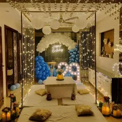 An intimate and cozy indoor birthday dinner setup. This features a cabana frame draped with fairy lights, low seating with plush cushions, and a balloon backdrop in shades of blue and white, perfect for a surprise celebration at home.