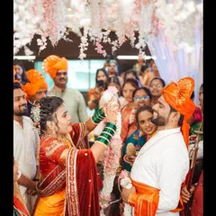 The bride joyfully places the garland on the groom, surrounded by smiling family and friends. This candid shot captures the festive atmosphere and collective happiness of the wedding ceremony.