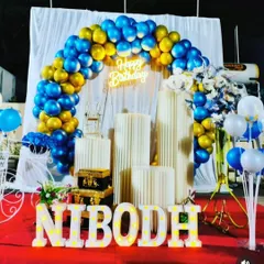 A striking blue and gold theme set against a white drape backdrop. This setup includes a balloon arch, neon sign, and light-up marquee letters spelling 'NIBODH' on a red carpet.