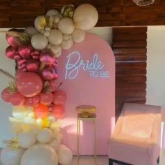 This modern "Bride to be" photo booth features a stylish pink arched panel with a custom neon sign. The look is completed with a cascading organic balloon garland in shades of pink, cream, and gold, perfect for creating memorable pictures.