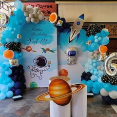 A fantastic space-themed birthday party for Ved. This decoration features custom backdrops with astronauts and planets, a rocket cutout, and a blue and orange balloon arrangement.
