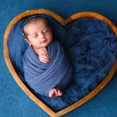 A beautiful composition featuring a newborn wrapped in deep blue fabric, nestled inside a wooden heart-shaped bowl. The rich color contrast creates a striking and lovely portrait.