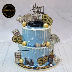 A luxurious two-tier birthday cake with a money and car theme. This 5kg black forest cake features a blue drip design, edible money, a toy jeep, and Ferrero Rocher chocolates.