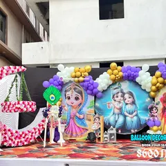A grand outdoor stage setup for a Krishna-themed birthday. The decor includes a large floral moon prop, custom backdrops, and character cutouts.