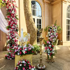 An outdoor decor setup for a traditional ceremony, featuring a floral arch of pink and white roses. The scene is accented with a classic brass lamp (diya) and a small elephant statue, blending sacred and celebratory elements.