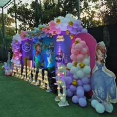 A dreamy Sofia the First princess party setup on a lush green lawn. The backdrop features the princess herself, a castle, and a beautiful balloon arch in shades of purple, pink, and gold.