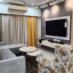 This living room combines comfort and style with a grey sectional sofa, a floral accent chair, and a modern TV unit. The design features a dark textured accent wall with track lighting, creating a focal point in this Thane apartment interior.
