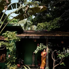 A bride stands on the porch of a green-walled suite, nestled amongst tropical plants. The vibrant colors make her traditional attire pop.