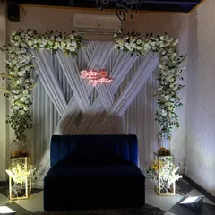 For modern engagement or roka ceremonies, I create clean and stylish backdrops. This setup uses white drapes, a floral arch, and a custom 'Better Together' neon sign for a chic photo corner.