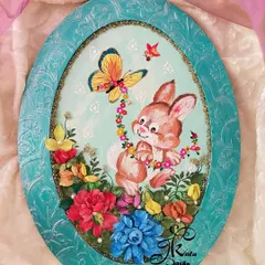 A throwback to an adorable oval wall panel featuring a bunny playing with a butterfly amidst a field of sculpted flowers. The textured blue frame adds a charming finish.