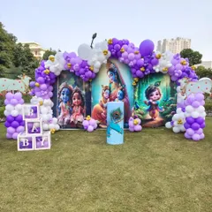 For this outdoor baby shower, we designed a beautiful and unique Krishna theme. The setup includes a large, vibrant backdrop with illustrations of Little Krishna, framed by a purple and white organic balloon arch, and custom 'BABY' blocks, all arranged perfectly on a lush green lawn.