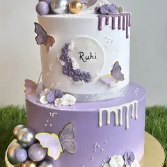 This elegant two-tiered birthday cake combines soft violet and white whipped cream frosting. We decorated it with delicate butterflies, chocolate drips, and shimmering spheres for a sophisticated and beautiful celebration cake.