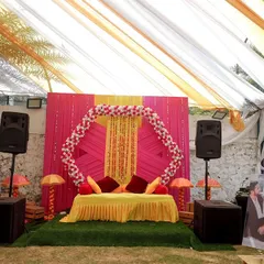 A bright and cheerful Haldi setup for a destination wedding. The decor features a pink and yellow backdrop with a hexagonal floral frame and personalized photos of the couple.