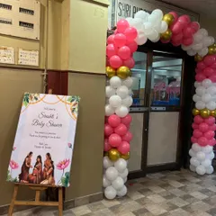 A simple and elegant entrance for a baby shower, featuring a welcome sign on an easel and a classic pink, white, and gold balloon arch.