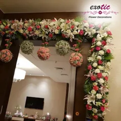 A lavish floral frame for a wide doorway, featuring white lilies, pink roses, and hanging floral balls. This full and abundant design makes a grand statement for weddings, engagements, or major festivals.