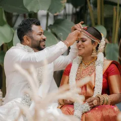 A candid moment from a morning wedding ceremony. The groom lovingly places the sindoor on the bride, with the lush green foliage providing a serene and natural background.