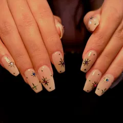 Subtle elegance with starry details. This beautiful manicure features a sheer nude base on long, coffin-shaped nails, adorned with tiny metallic stars, colorful rhinestones, and studs for a celestial look.