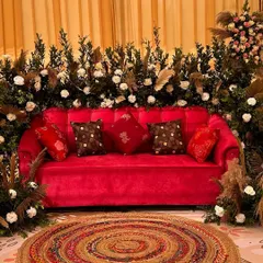A pop of color in a boho setting. A vibrant red velvet sofa is surrounded by an installation of pampas grass and white florals, with a colorful jute rug to complete the look.